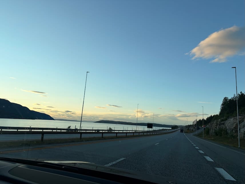 An image taken from the passenger seat of a car. There is a long road stretching into the distance, with hills and a large lake to the side. The sky is clear and blue.