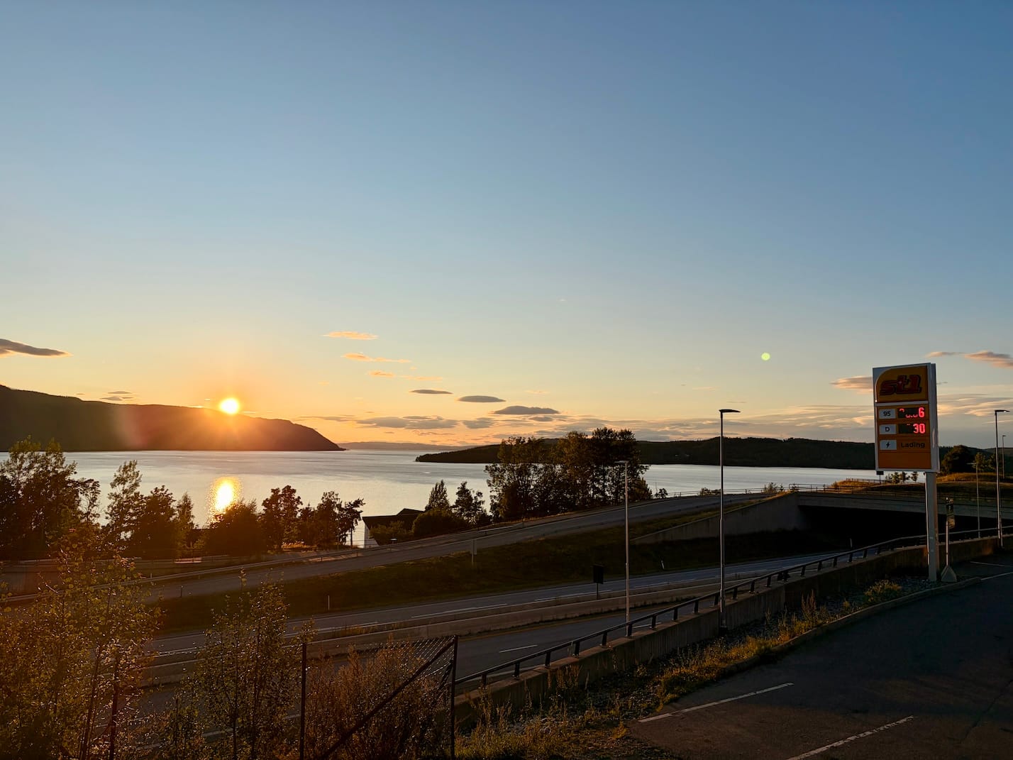 An image of a motorway in Norway. There's a lake and a setting sun in the background.